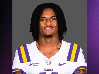 Portrait of an LSU football player with dreadlocks, wearing a white jersey with purple and gold stripes, against a gray backdrop.
