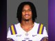 Portrait of an LSU football player with dreadlocks, wearing a white jersey with purple and gold stripes, against a gray backdrop.