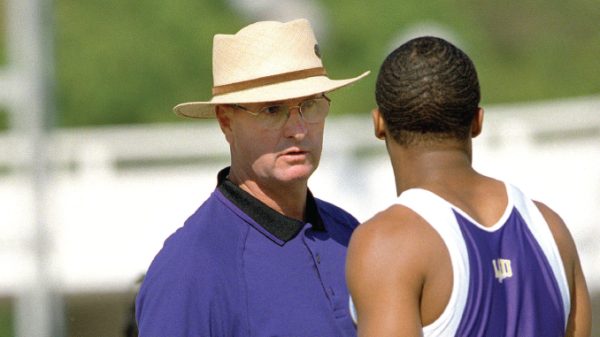 Coach wearing a straw hat talks to a sprinter in a purple singlet on a track outside.