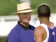 Coach wearing a straw hat talks to a sprinter in a purple singlet on a track outside.