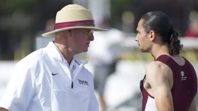 Coach in a white jacket and straw hat talks to a male athlete in a maroon and white track uniform on a blurred outdoor track field.