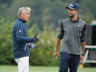 Two coaches converse on a grassy outdoor practice field, one gesturing with gloved hands left of center.