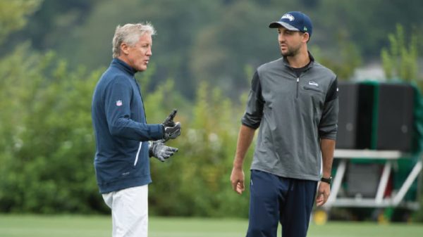 Two coaches converse on a grassy outdoor practice field, one gesturing with gloved hands left of center.