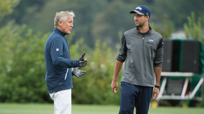 Two coaches converse on a grassy outdoor practice field, one gesturing with gloved hands left of center.