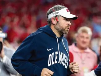 Man in an Ole Miss hoodie and white visor running on the football sidelines during a game.