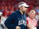 Man in an Ole Miss hoodie and white visor running on the football sidelines during a game.