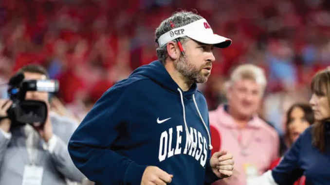 Man in an Ole Miss hoodie and white visor running on the football sidelines during a game.