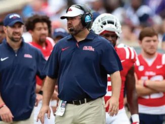 Football coach on the sideline wearing a headset and navy Ole Miss polo, khaki pants, and an ID badge, with players in red jerseys behind him.