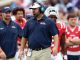 Football coach on the sideline wearing a headset and navy Ole Miss polo, khaki pants, and an ID badge, with players in red jerseys behind him.