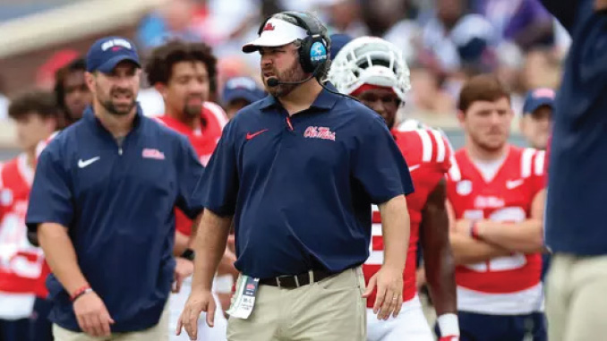 Football coach on the sideline wearing a headset and navy Ole Miss polo, khaki pants, and an ID badge, with players in red jerseys behind him.