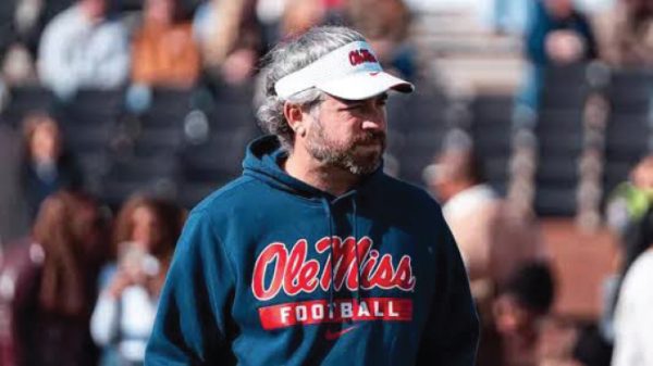 Side-profile of a man wearing a blue Ole Miss Football hoodie and a white visor at a stadium, looking to the right with a crowd behind him.