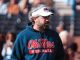 Side-profile of a man wearing a blue Ole Miss Football hoodie and a white visor at a stadium, looking to the right with a crowd behind him.