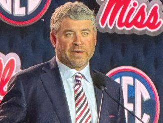 Man in a dark suit and striped tie speaks at a podium with Miss logos in the background.