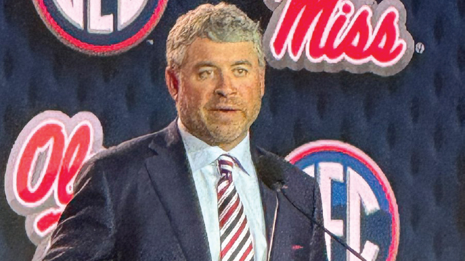 Man in a dark suit and striped tie speaks at a podium with Miss logos in the background.