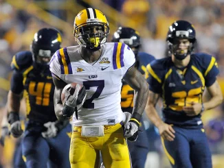 LSU football player in white and purple, wearing #7, sprints with the ball as defenders in navy and gold chase him on the field.