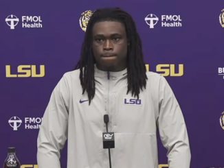 LSU football player at a press conference podium wearing a light gray LSU jacket with a purple backdrop and LSU logos behind him.