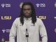 LSU football player at a press conference podium wearing a light gray LSU jacket with a purple backdrop and LSU logos behind him.