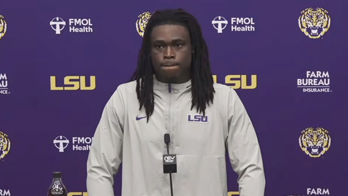 LSU football player at a press conference podium wearing a light gray LSU jacket with a purple backdrop and LSU logos behind him.