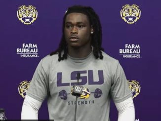 African American LSU football player at a podium during a press conference, wearing a gray LSU Strength shirt with a purple backdrop of logos.
