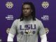 African American LSU football player at a podium during a press conference, wearing a gray LSU Strength shirt with a purple backdrop of logos.