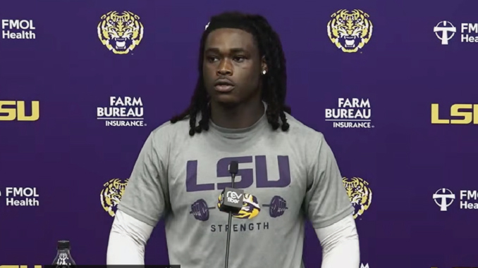 African American LSU football player at a podium during a press conference, wearing a gray LSU Strength shirt with a purple backdrop of logos.