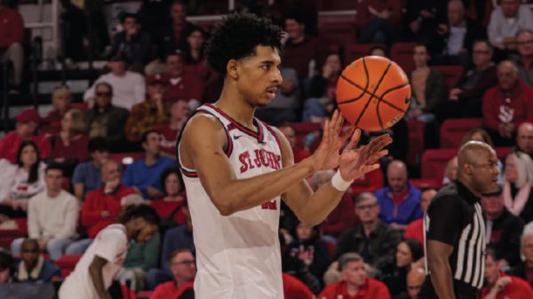 Basketball player in a white St. Joseph jersey passes the ball during a game, with spectators in the stands in the background.