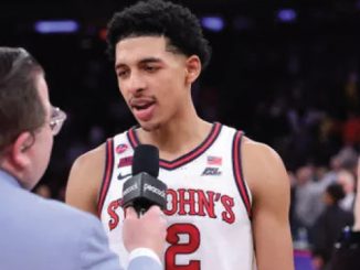 Basketball player in a St. John's jersey being interviewed on court, microphone held near his mouth, crowd in the background, jersey number 2 visible.