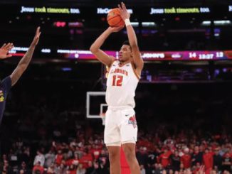 College basketball player in white jersey number 12 rises for a jump shot while defender in navy, number 5, guards him; arena filled with fans is in the background.