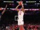College basketball player in white jersey number 12 rises for a jump shot while defender in navy, number 5, guards him; arena filled with fans is in the background.