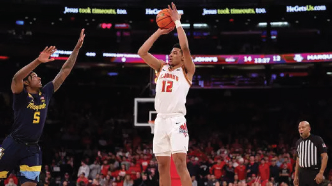 College basketball player in white jersey number 12 rises for a jump shot while defender in navy, number 5, guards him; arena filled with fans is in the background.