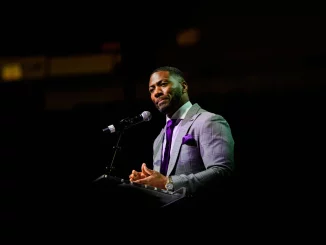 Man in a gray checkered suit and purple tie speaks at a podium with microphone on a dark stage.