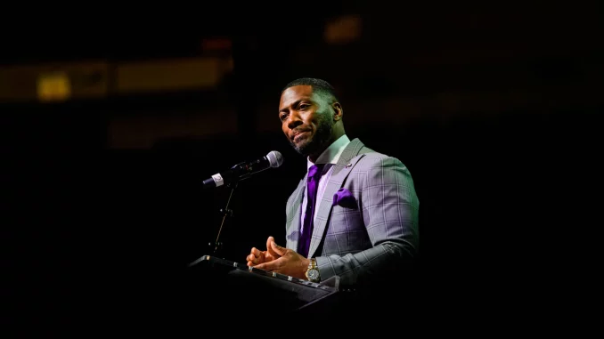 Man in a gray checkered suit and purple tie speaks at a podium with microphone on a dark stage.