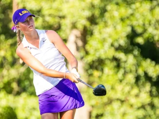 Female golfer mid-swing wearing a purple LSU cap and white polo on a sunny golf course.