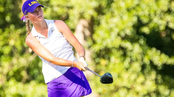 Female golfer mid-swing wearing a purple LSU cap and white polo on a sunny golf course.