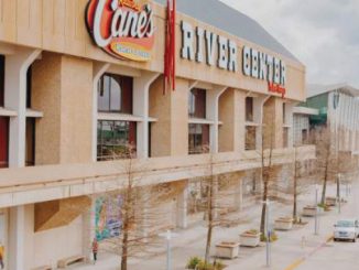 Exterior view of River Center mall with a Raising Cane's sign and a row of bare trees along the sidewalk