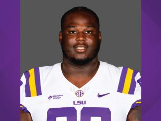Portrait of a football player wearing a white LSU jersey with purple and gold accents, facing the camera against a gray backdrop.
