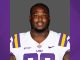 Portrait of a football player wearing a white LSU jersey with purple and gold accents, facing the camera against a gray backdrop.
