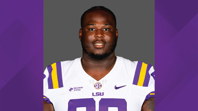 Portrait of a football player wearing a white LSU jersey with purple and gold accents, facing the camera against a gray backdrop.