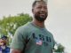 Smiling man with a beard wearing a green U.S. shirt and sunglasses on his head, outdoors on a sunny day.