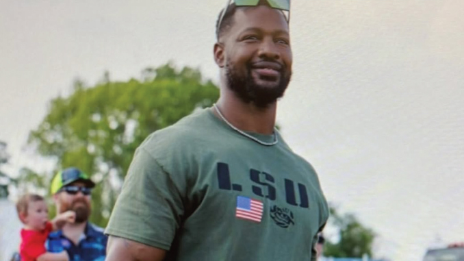 Smiling man with a beard wearing a green U.S. shirt and sunglasses on his head, outdoors on a sunny day.
