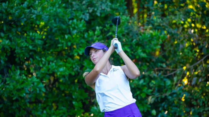 Female golfer in a white polo and purple shorts mid-swing with a golf club, against a leafy green backdrop.