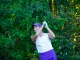 Female golfer in a white polo and purple shorts mid-swing with a golf club, against a leafy green backdrop.