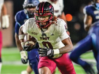 College football running back in a white Troy jersey with the ball, sprinting downfield past defenders biology-blurred background