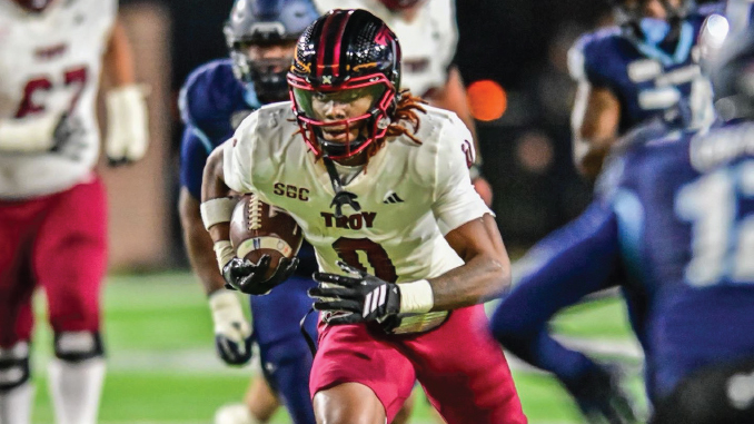 College football running back in a white Troy jersey with the ball, sprinting downfield past defenders biology-blurred background