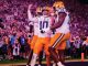 LSU football players in white jerseys celebrate on the field as the stadium cheers in the background.