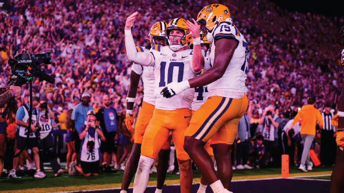 LSU football players in white jerseys celebrate on the field as the stadium cheers in the background.