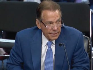Man in a blue suit and glasses testifying at a hearing, speaking into a microphone in a formal setting.