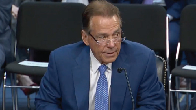 Man in a blue suit and glasses testifying at a hearing, speaking into a microphone in a formal setting.