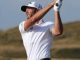 Male golfer in a white polo shirt and cap mid-swing on a golf course, follow-through with a raised club.