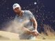 Male golfer in a white Callaway cap executes a bunker shot, sand spraying as he swings with his club.
